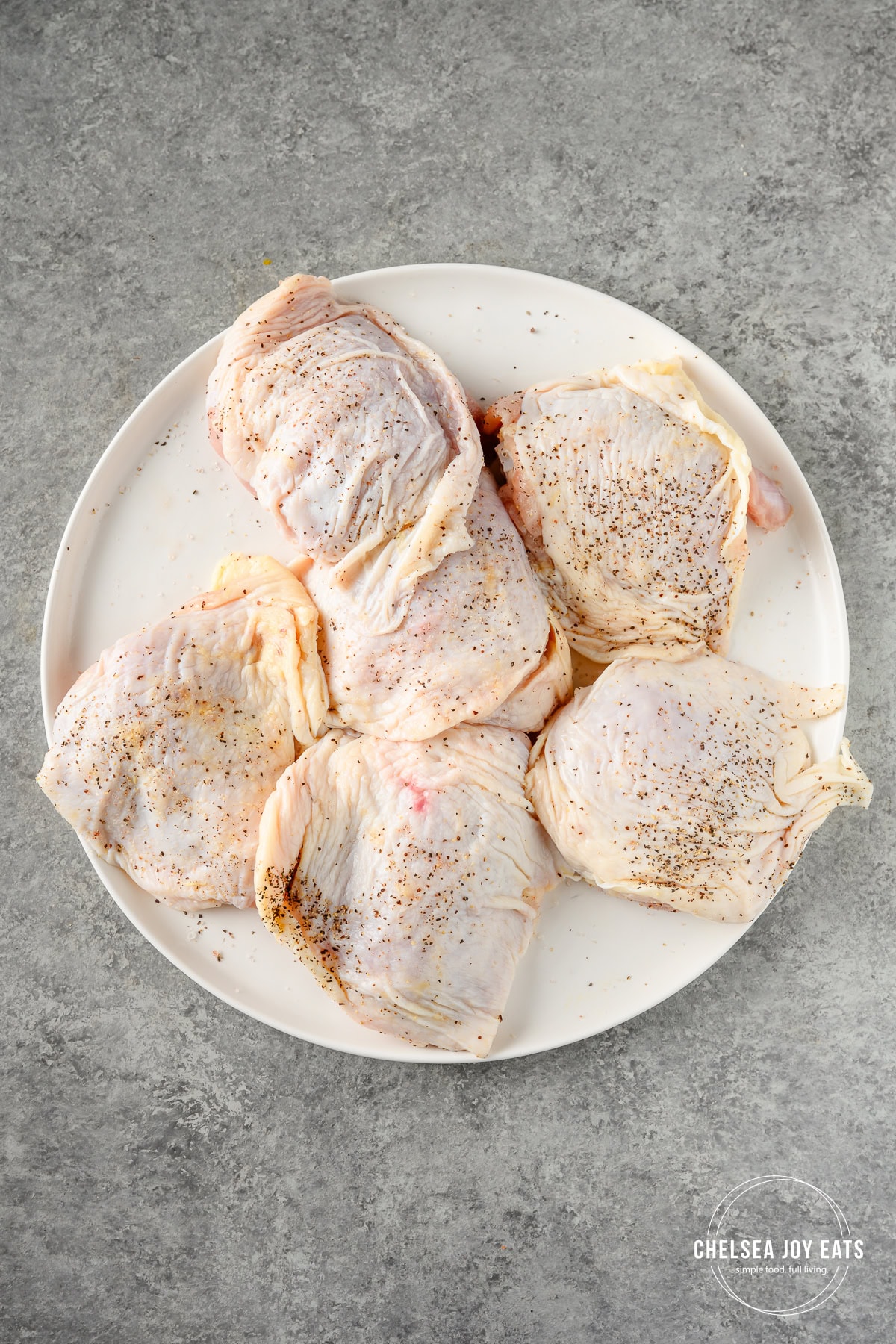 Uncooked bone-in, skin-on chicken thighs seasoned with salt and pepper on a white plate.