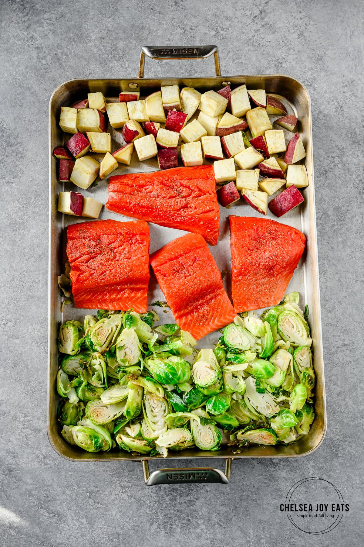 Sheet pan with partially roasted sweet potatoes and brussels sprouts with raw salmon fillets added to the middle.
