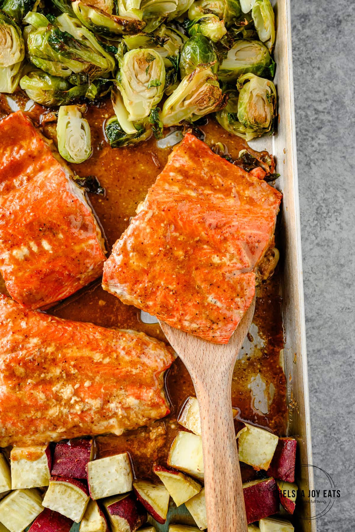Roasted maple mustard salmon with brussels sprouts and sweet potatoes on a sheet pan with a spatula lifting a portion of salmon.