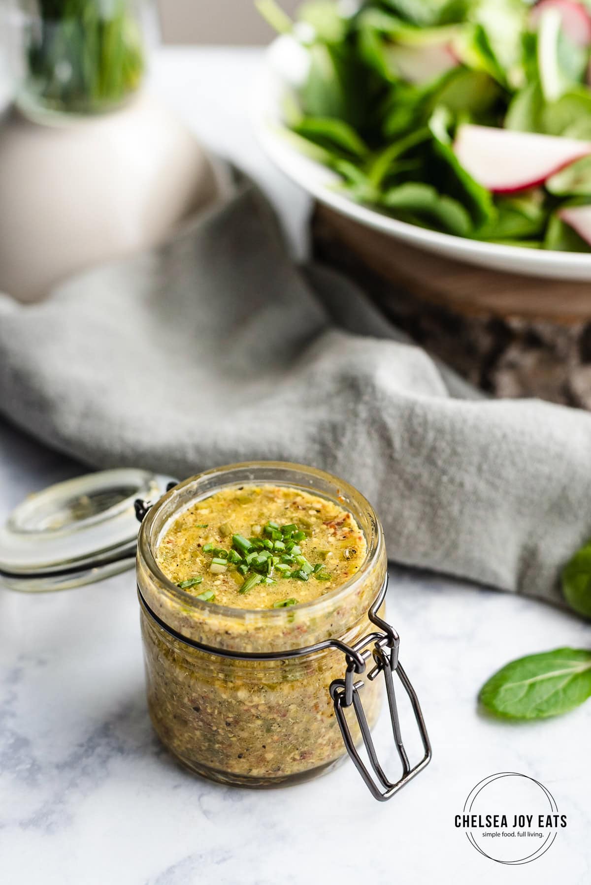 Small glass jar filled with mustard chive vinaigrette with a salad and linens in the background.
