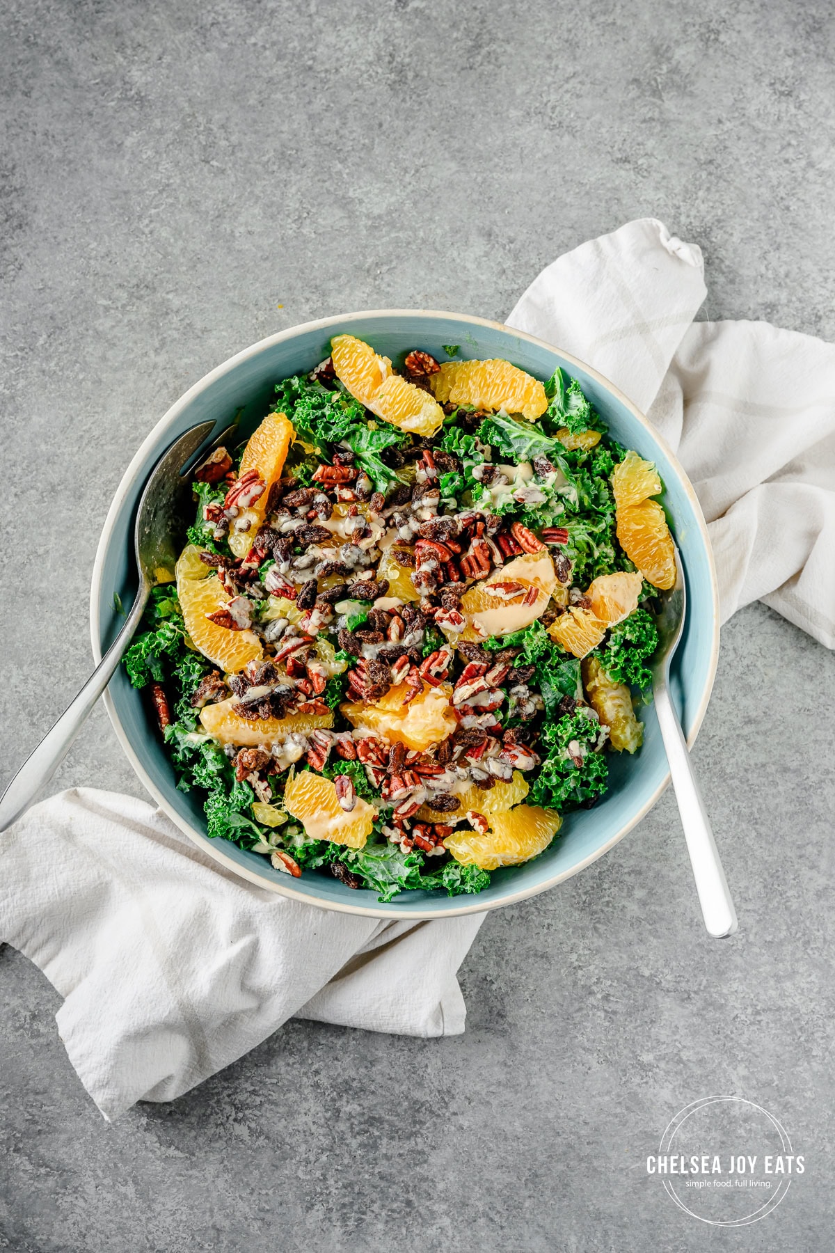 Oranges, pecans, and raisins on a bed of kale drizzled with citrus tahini dressing in a serving bowl with serving spoons alongside.