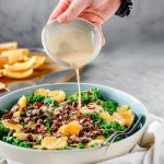 Person pouring citrus tahini dressing over a Orange Kale Salad with Pecans and Raisins, with cutting board of sliced oranges behind.