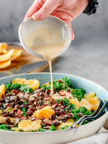Person pouring citrus tahini dressing over a Orange Kale Salad with Pecans and Raisins, with cutting board of sliced oranges behind.