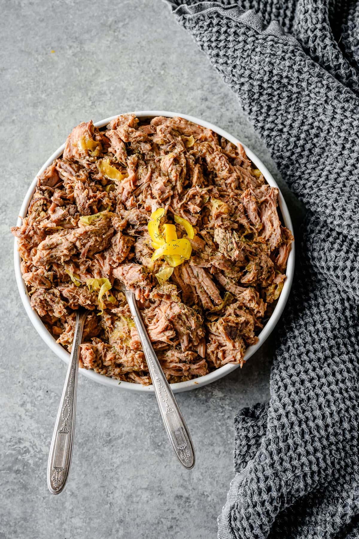 Bowl with shredded mississippi pot roast garnished with pepperoncini and ready with two serving spoons.