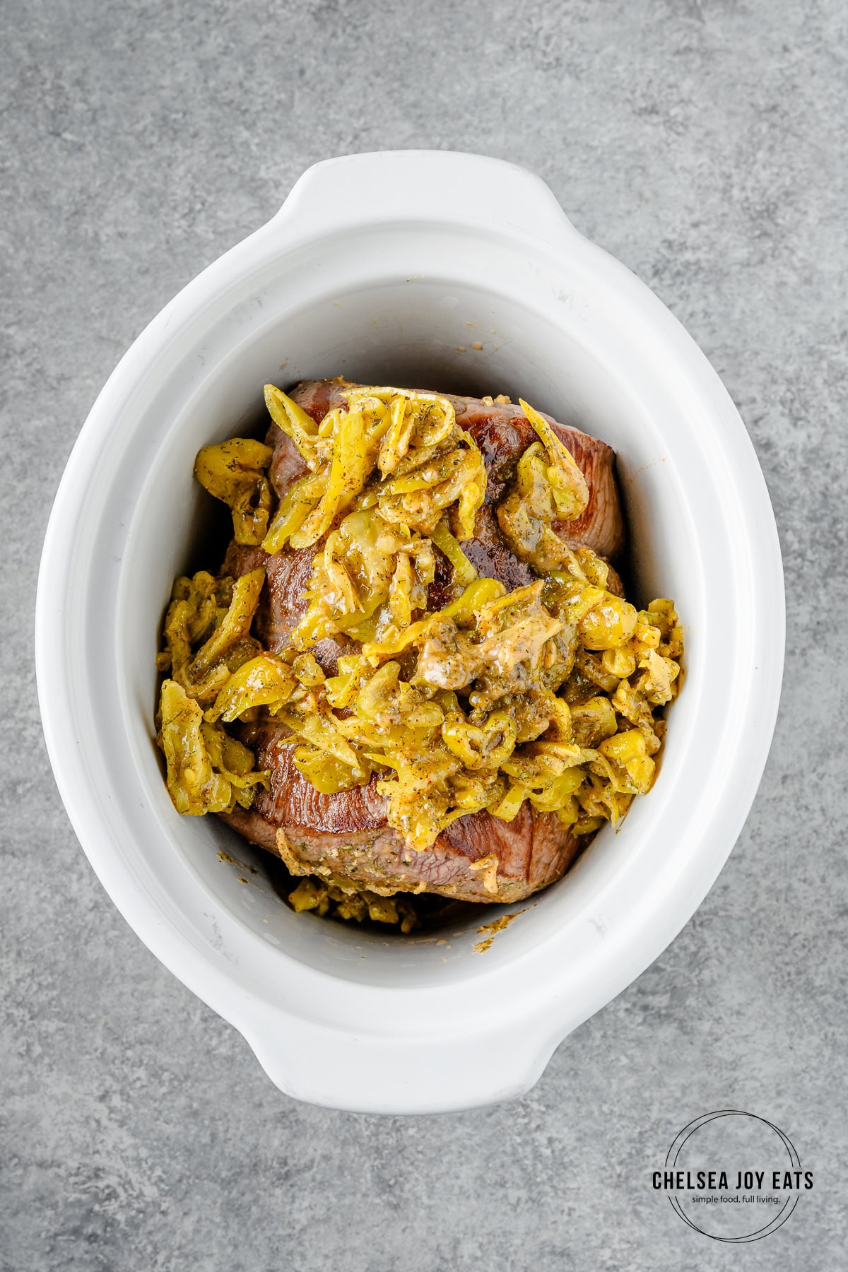 Beef roast in the crock of a slow cooker with a pepperoncini and spice mixture poured overtop.