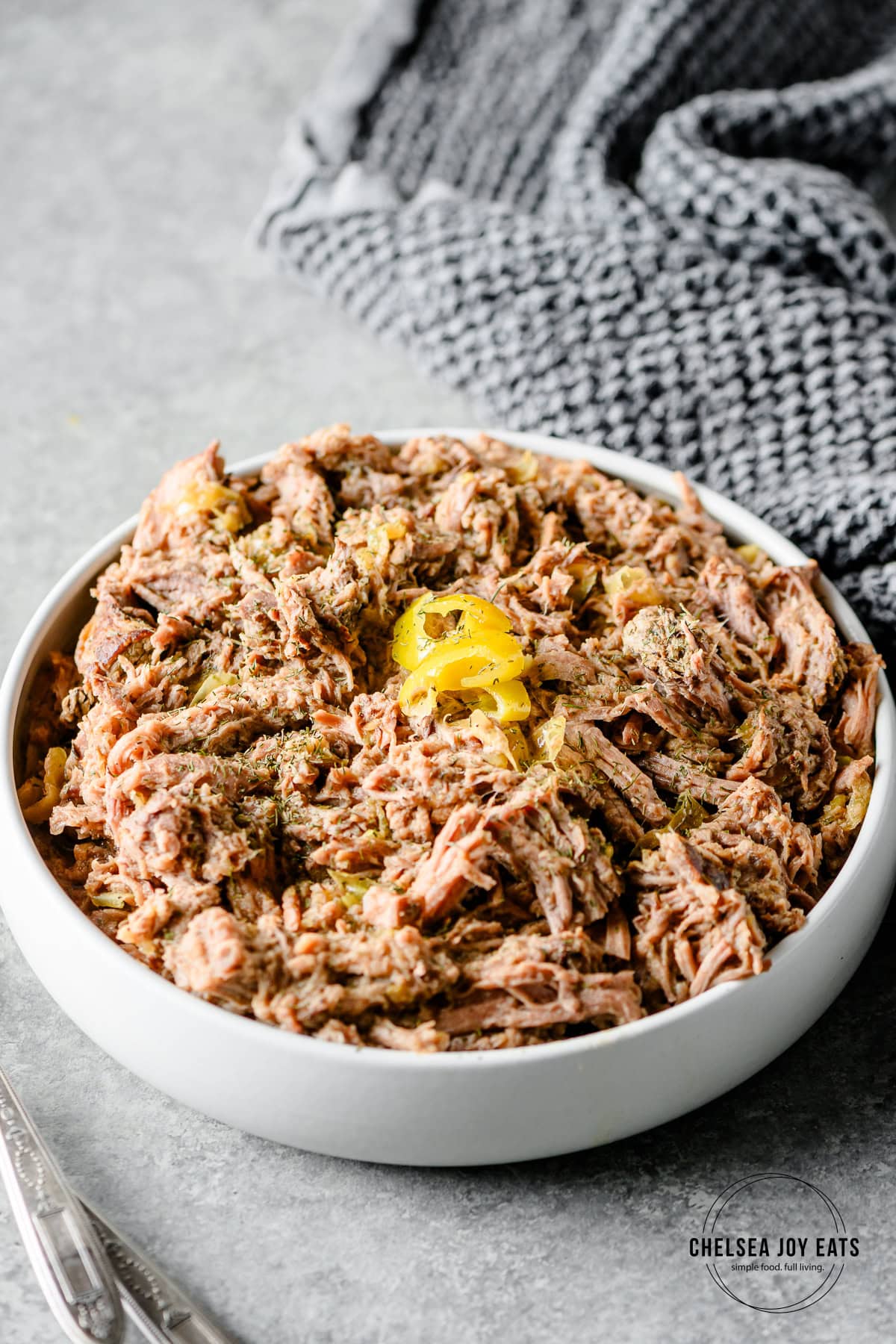 Bowl with shredded pepperoncini beef roast, topped with pepperoncini and dried dill, served alongside linens and serving spoons.