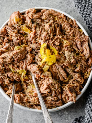 Bowl filled with pepperoncini shredded beef, garnished with pepperoncinis and dill, with two serving spoons.