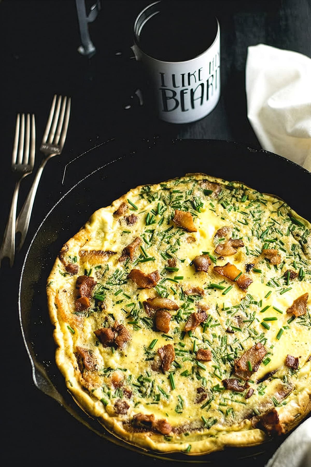 Plantain frittata with bacon and chives in a cast iron skillet on a dark table with forks and a mug of coffee alongside.
