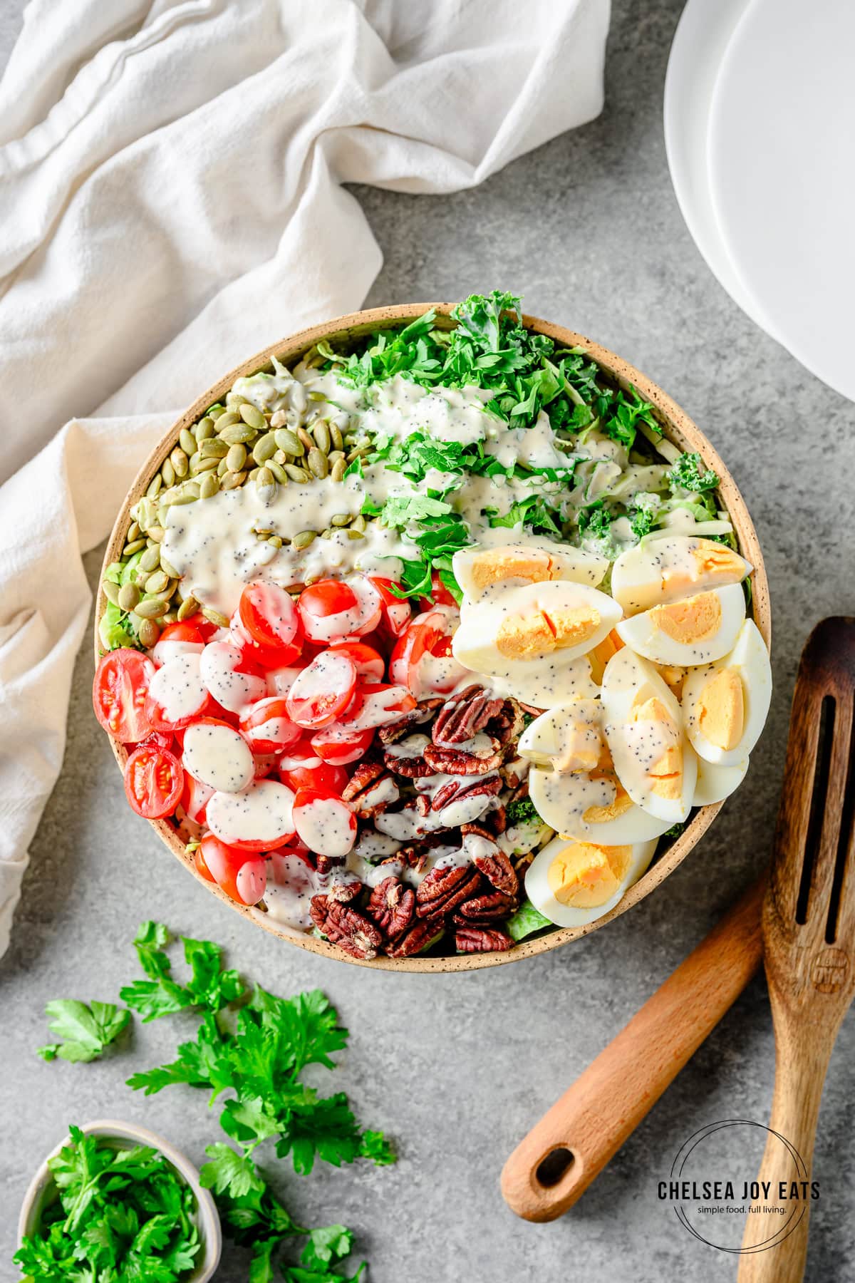 Chopped kale salad with brussels sprouts, hard-boiled eggs, nuts, and tomatoes in a large bowl with linens and herbs besides.