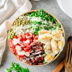 Chopped kale salad with brussels sprouts, hard-boiled eggs, nuts, and tomatoes in a large bowl with linens and herbs besides.