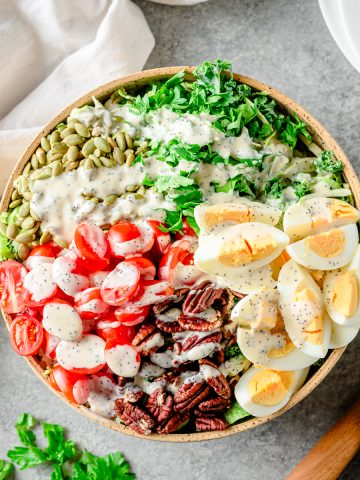Chopped kale salad with brussels sprouts, hard-boiled eggs, nuts, and tomatoes in a large bowl with linens and herbs besides.