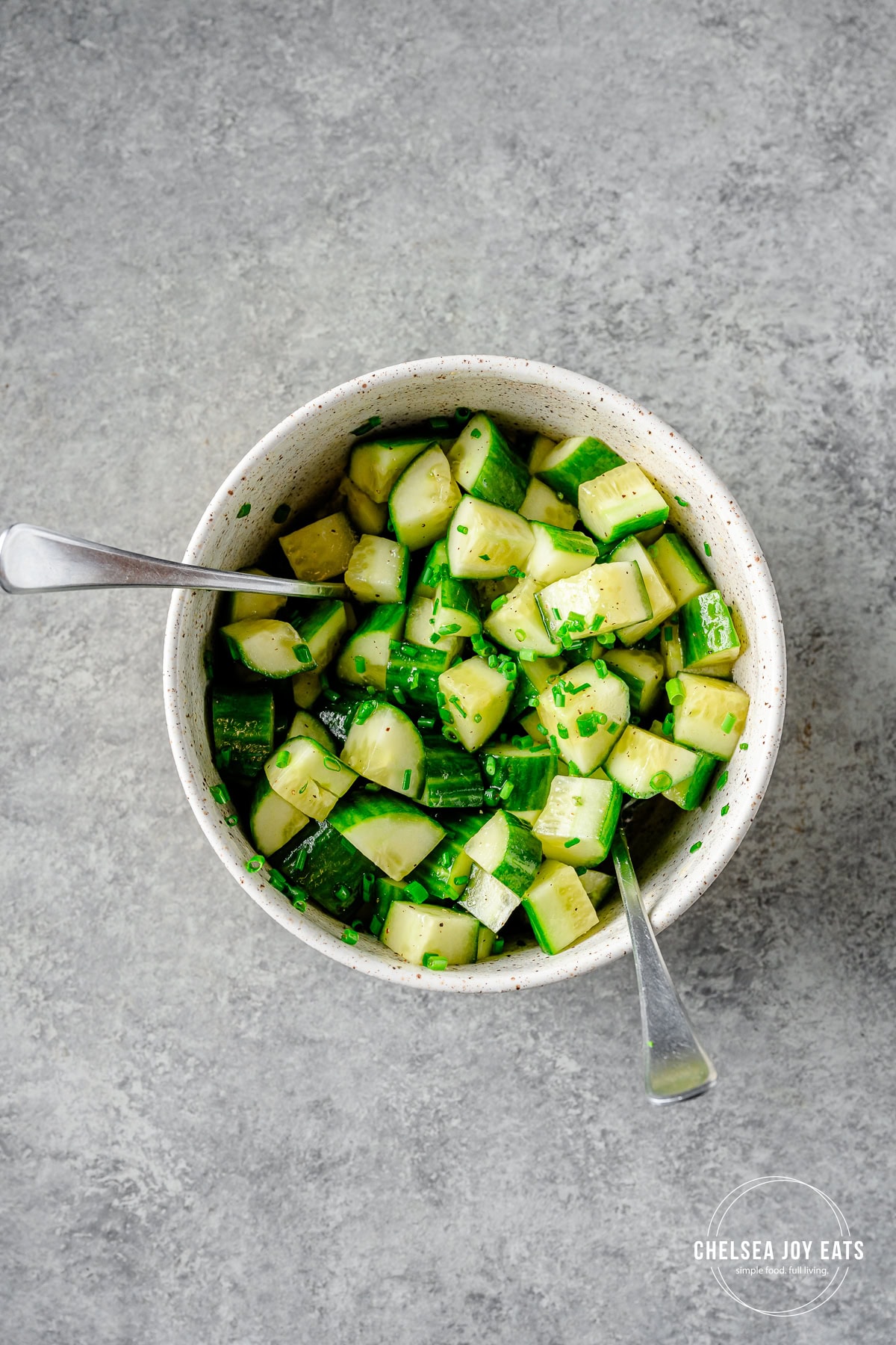 Cubed cucumbers tossed with chives and apple cider vinaigrette in a ceramic bowl with two forks.
