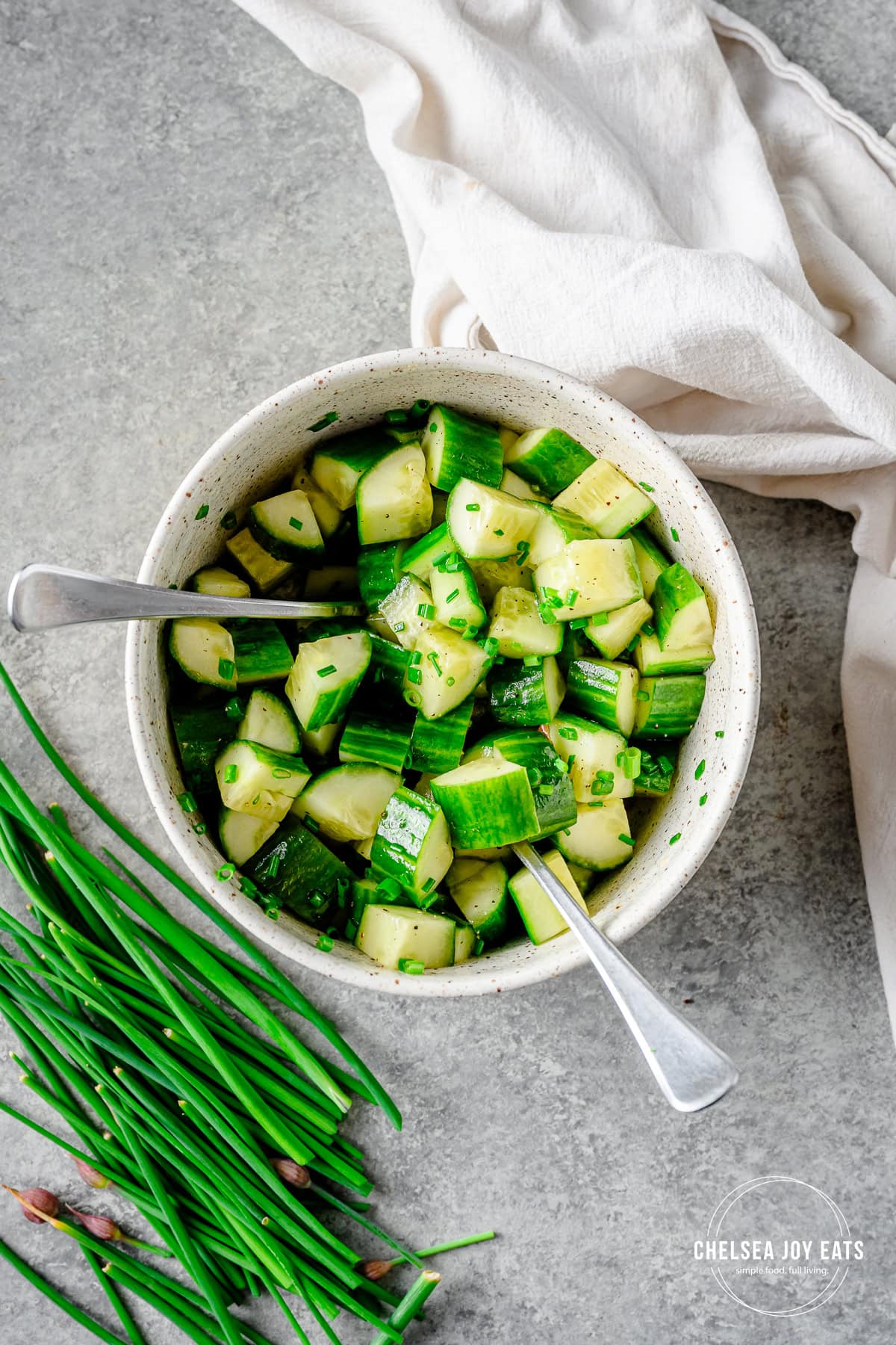 Chopped cucumber salad with chives in a pottery bowl with two forks, with white linens and fresh chives in the background.
