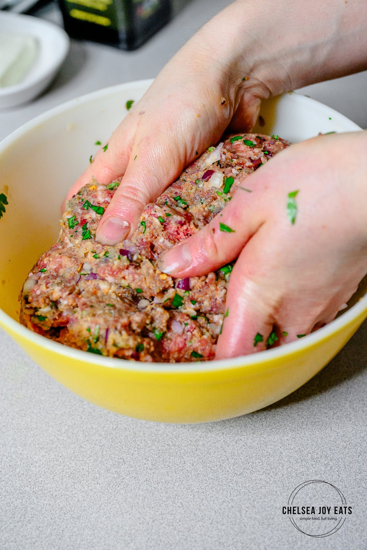 Hands mixing together leamb and beef in a mixing bowl with parsley, onions, and more spices.