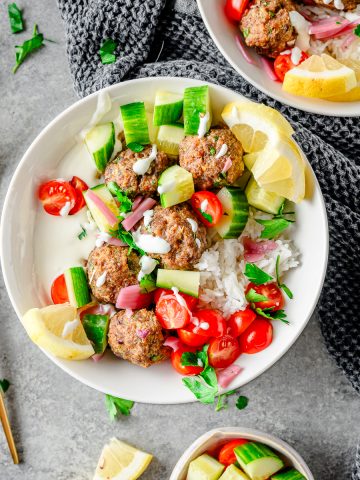 Bowl with rice, whipped feta, beef and lamb meatballs, and tomatoes and cucumber with additional bowls and forks besides.