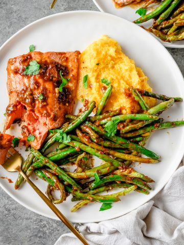 Plate with ginger miso salmon, polenta, and roasted green beans with a fork taking a bite out of the salmon.