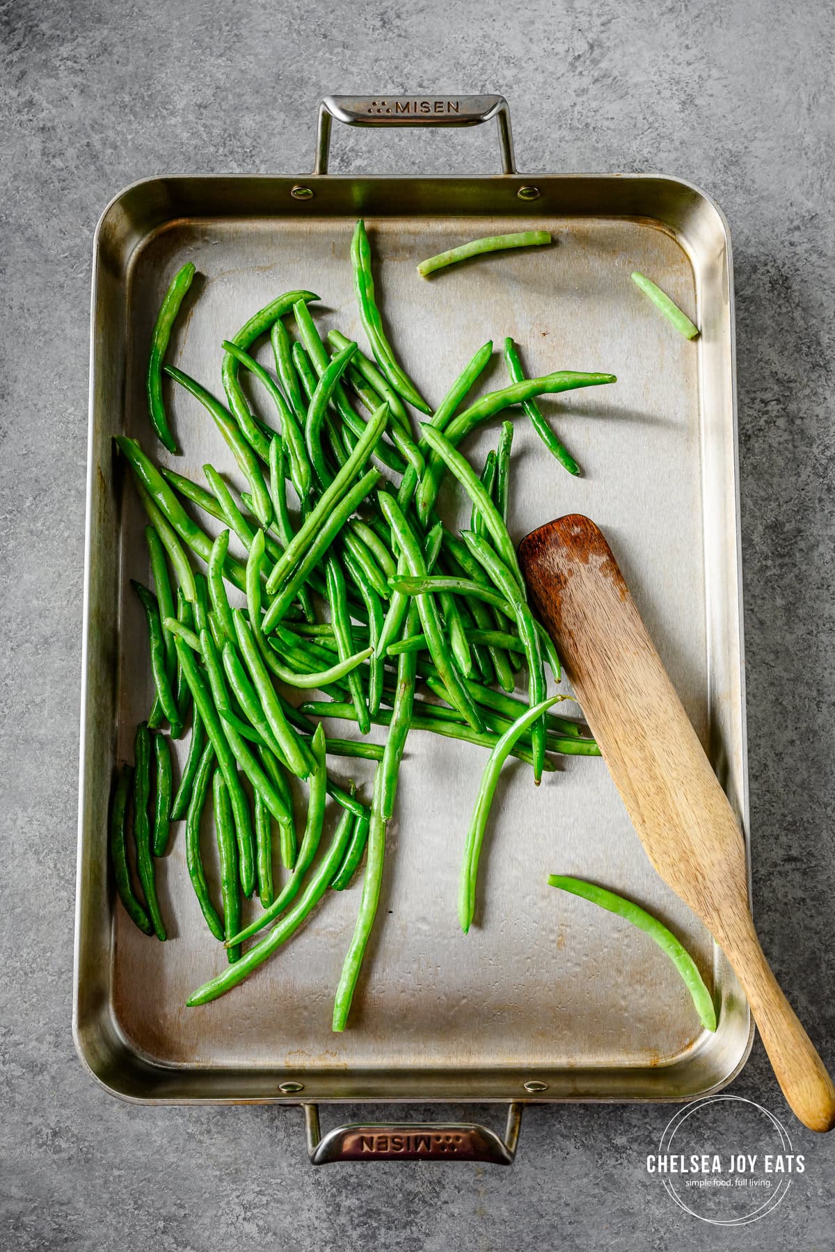 Green beans tossed in oil on a sheet pan with a wooden spatula.