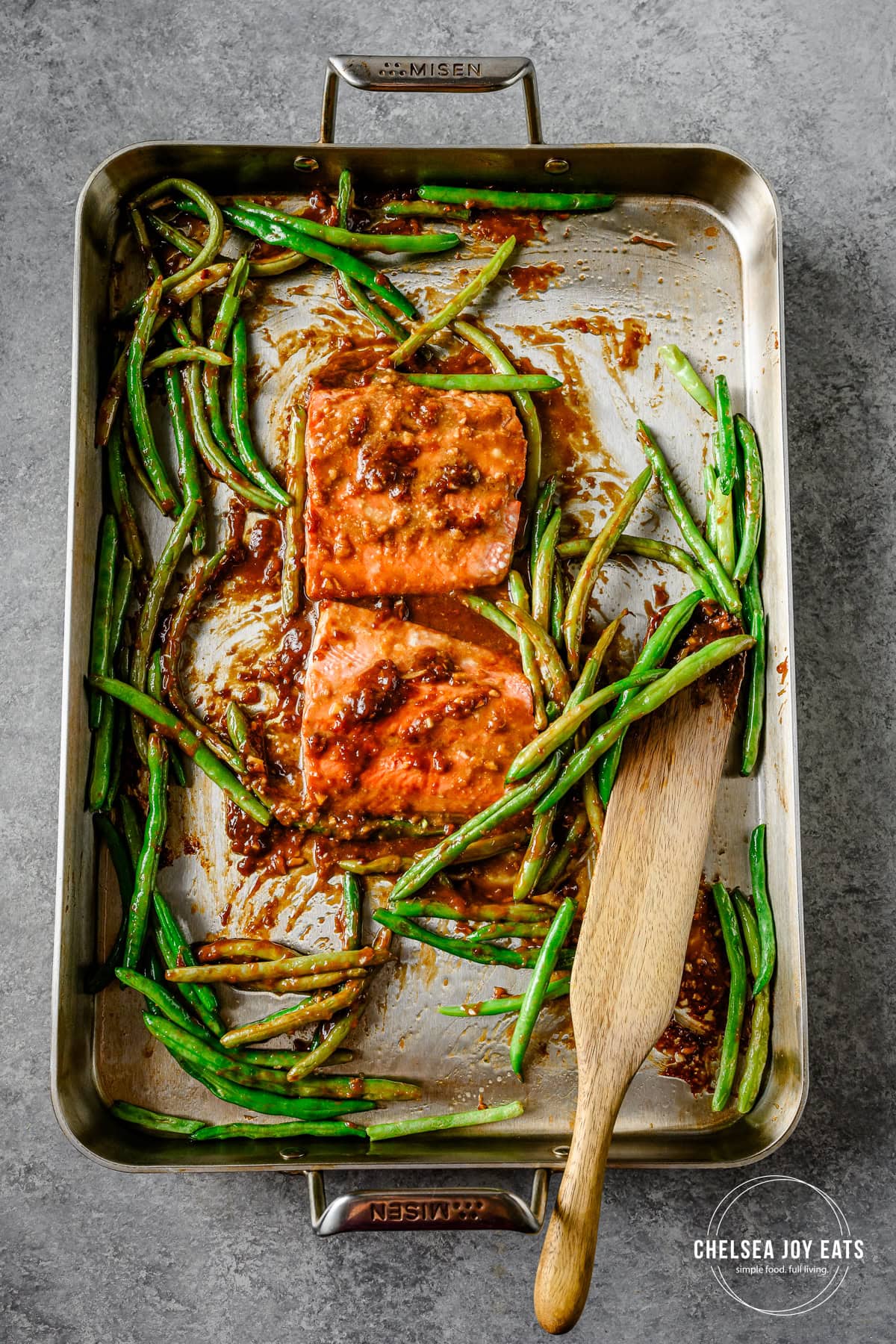 Roasted gluten free ginger miso salmon and green beans on a sheet pan, stirred with a wooden spatula.