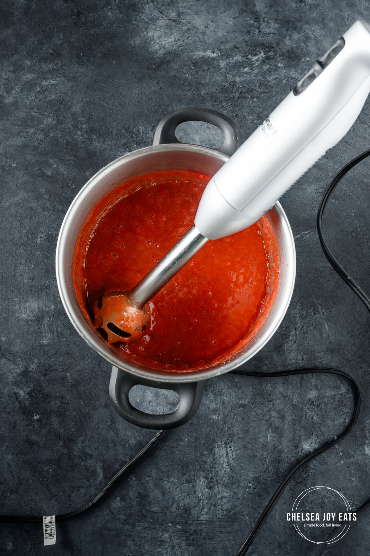 Immersion blender blending up cooked strawberries and rhubarb in a deep pot.