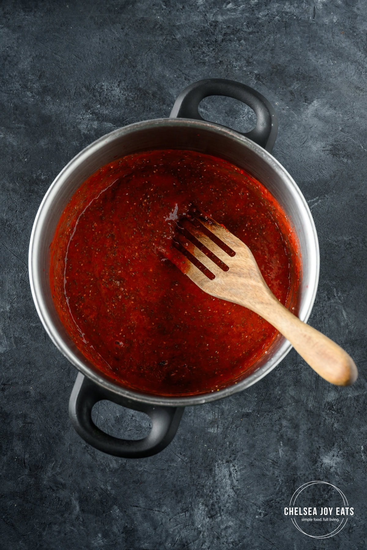 Strawberries and rhubarb puree mixed with chia seeds in a deep pot with a wooden spatula.
