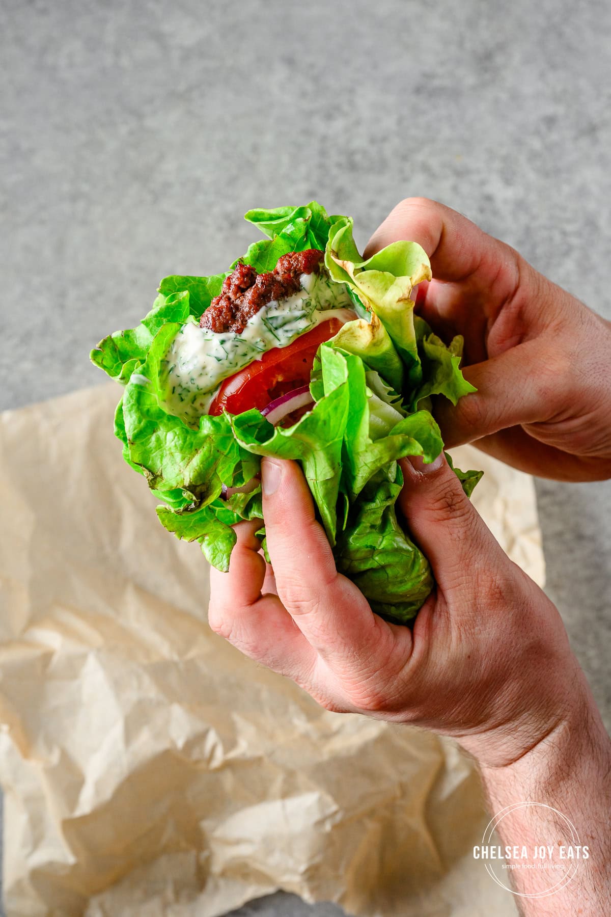 Lettuce-wrapped bunless burger with tomatoes, onion, pickles, and mayo sauce held up above a piece of parchment paper.