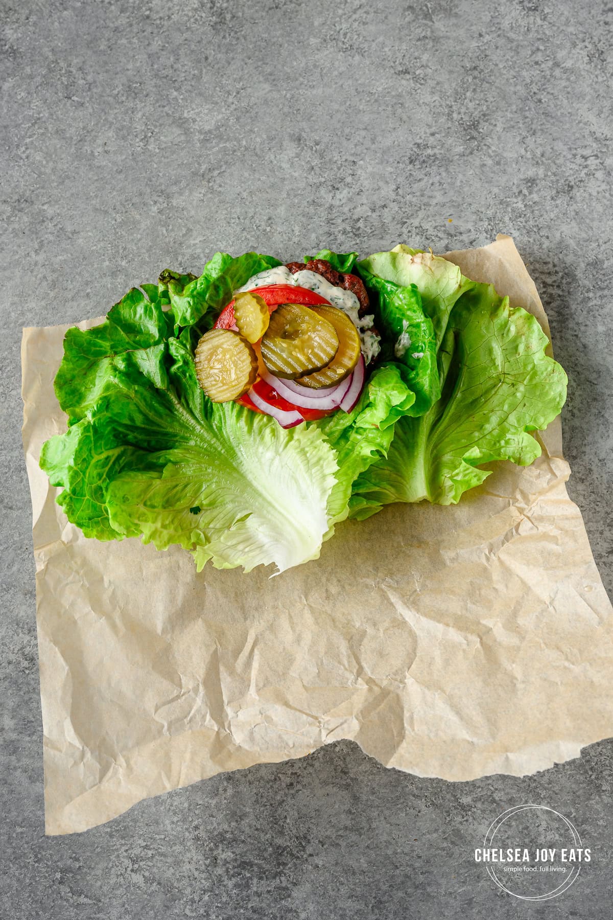 Bunless burger with onions, pickles, and onion on leaves of green lettuce with parchment paper.