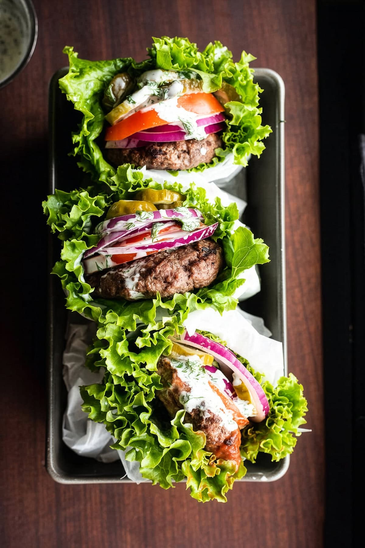 Lettuce-wrapped bunless burgers with tomato, onion, pickes, and cilantro-dill mayo sauce stacked in a loaf pan.