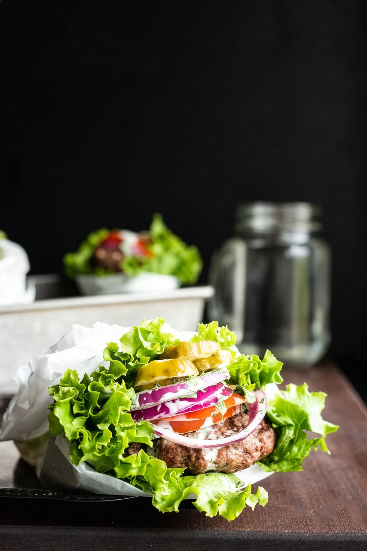 Lettuce-wrapped bunless burger topped with onion, tomato, pickles, and mayo sauce on a cutting board.