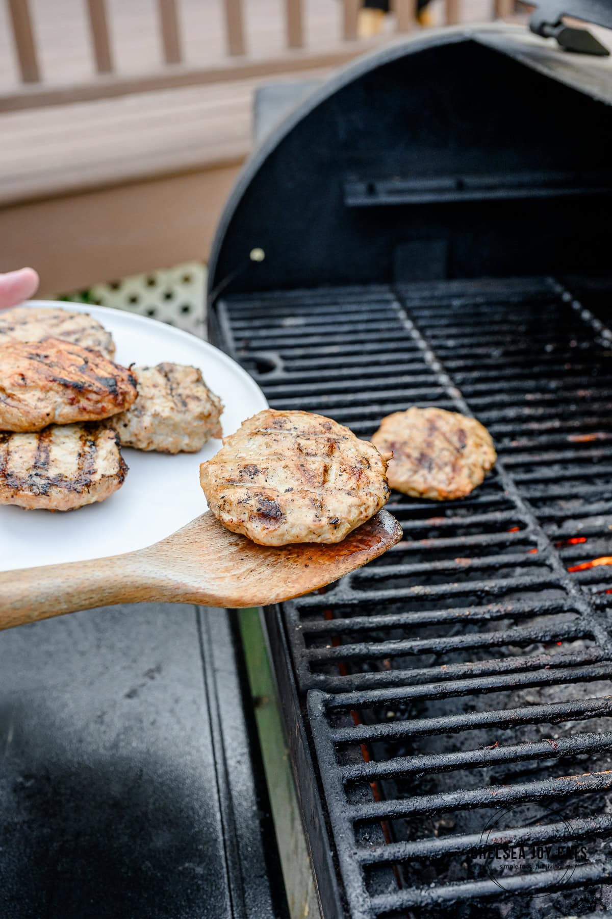 Person using a wooden spatula to take grilled chicken burger patties off of a grill and onto a white plate.