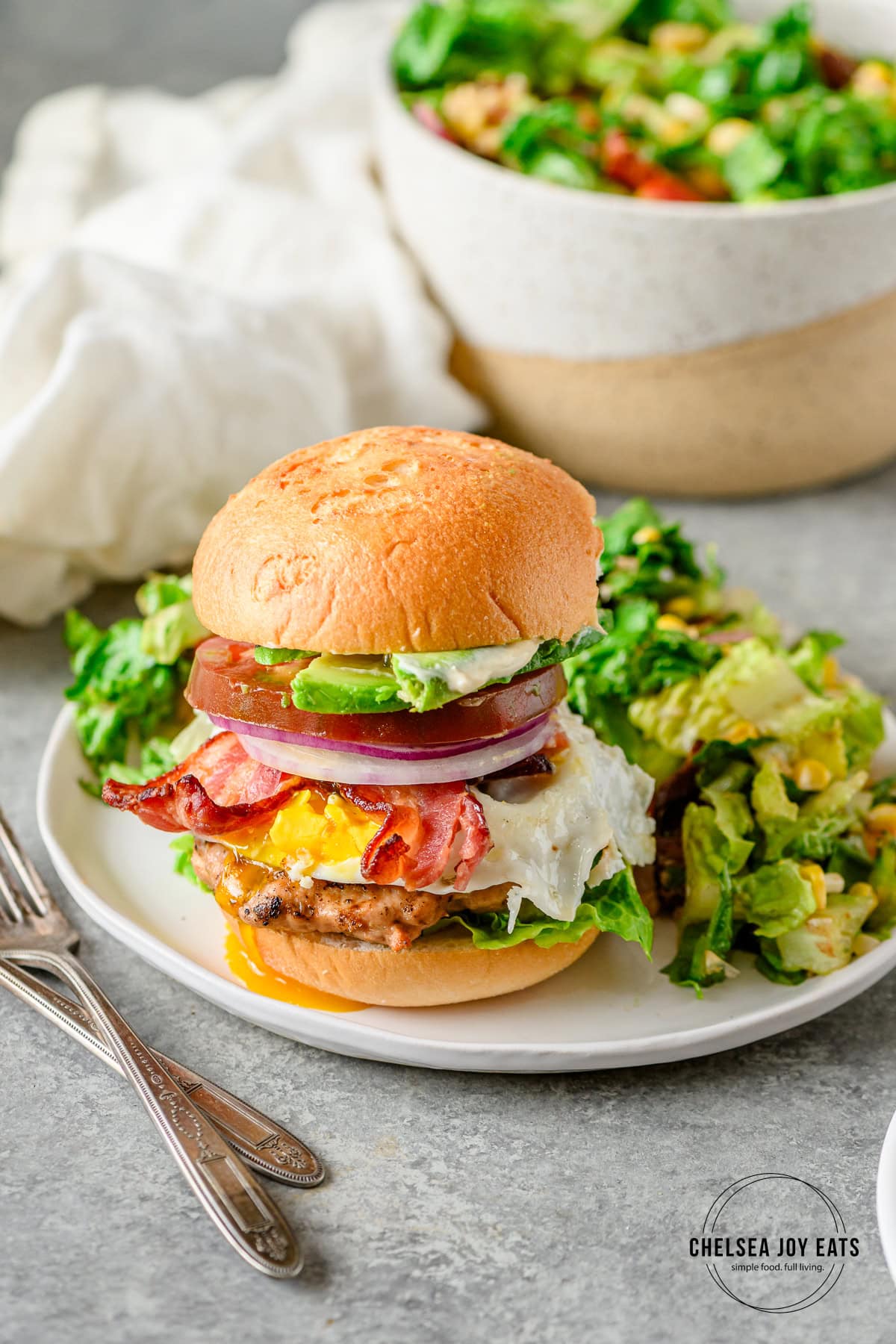 Gluten free chicken burger with toppings like egg, bacon, avocado, tomato, and mayonaisse on a gluten free bun served with a salad.