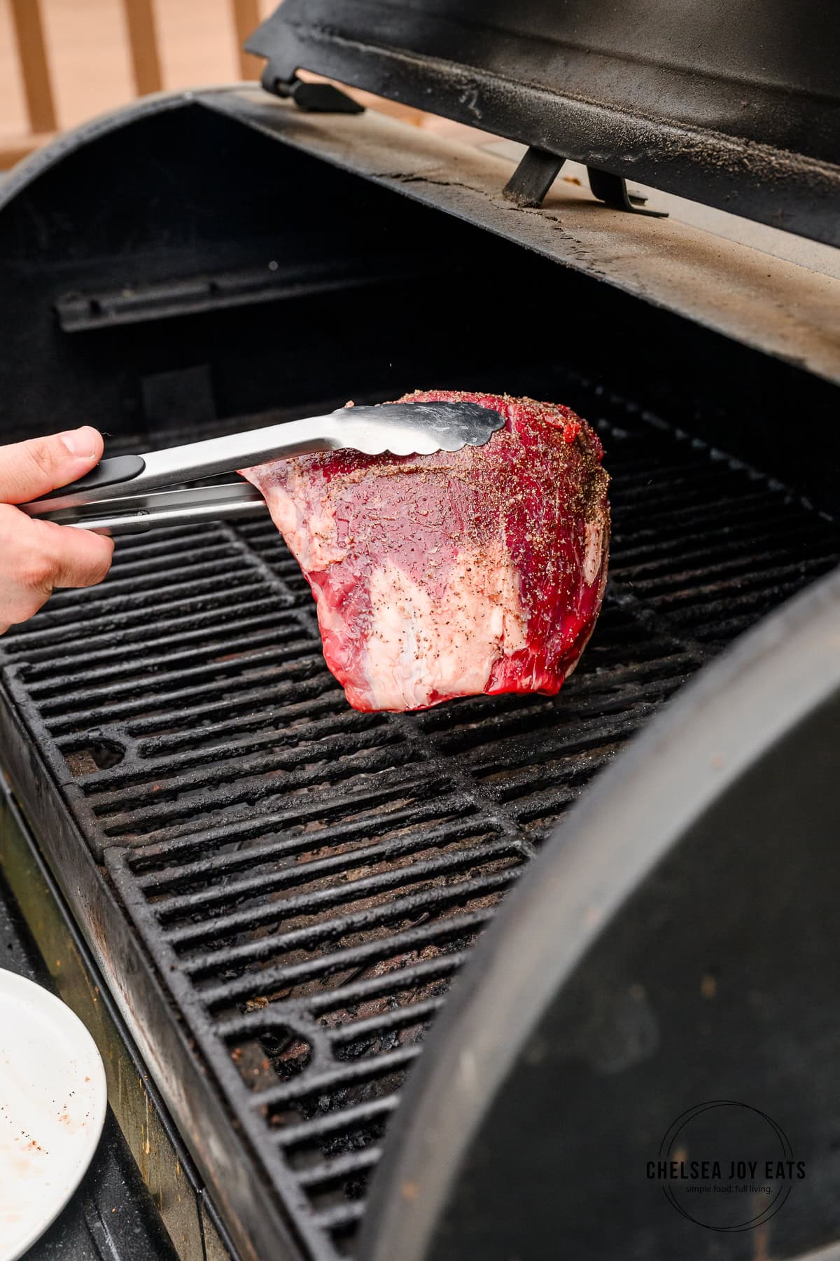Seasoned flank steak being placed on the grates of a grill with stainless steel tongs.