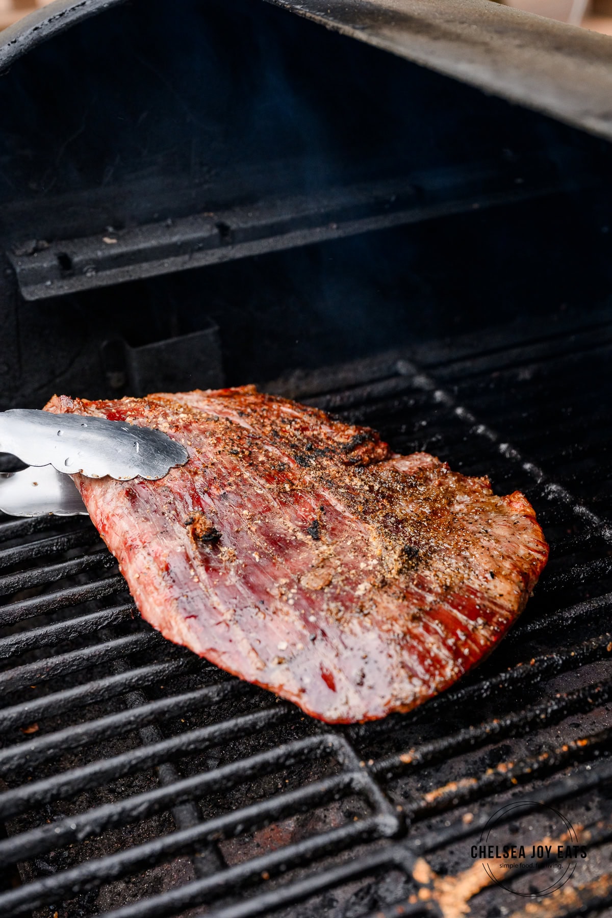 Flank steak being flipped with stainless steel tongs on a grill.