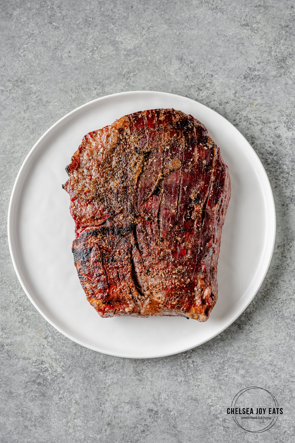 Grilled flank steak resting after cooking on a white plate on a grey countertop.