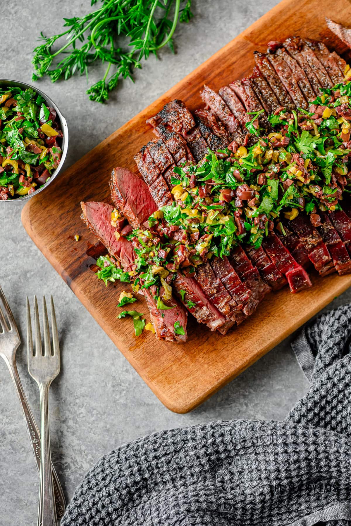 Sliced grilled flank steak topped with an olive herb relish on a wooden cutting board, with forks and grey linens besides.