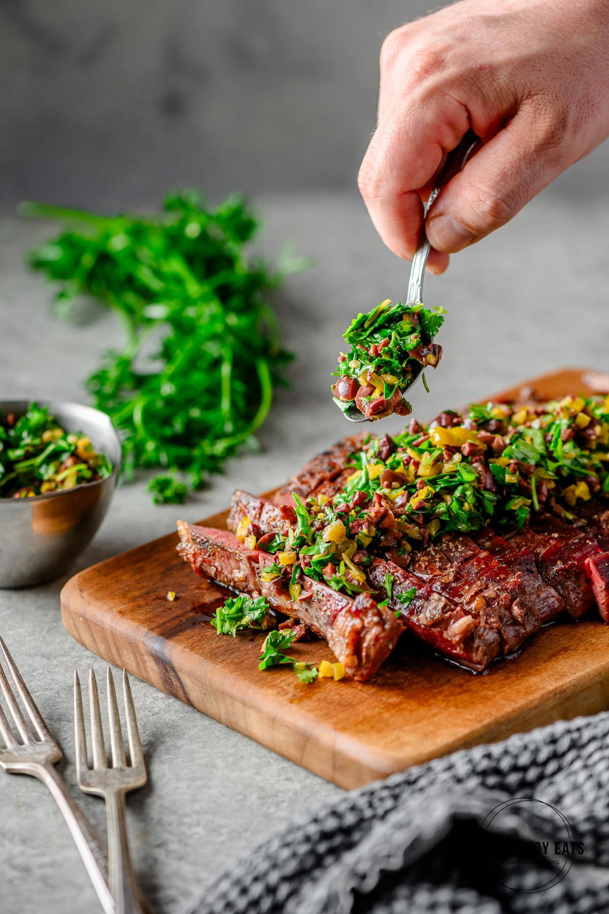 Sliced grilled flank steak on a woodern cutting board being topped with an olive herb relish, with a dish of relish and fresh parsley in the background.