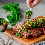 Sliced grilled flank steak on a woodern cutting board being topped with an olive herb relish, with a dish of relish and fresh parsley in the background.