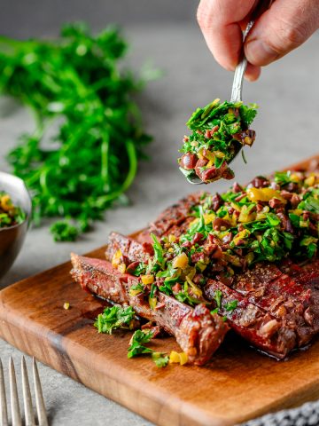 Sliced grilled flank steak on a woodern cutting board being topped with an olive herb relish, with a dish of relish and fresh parsley in the background.