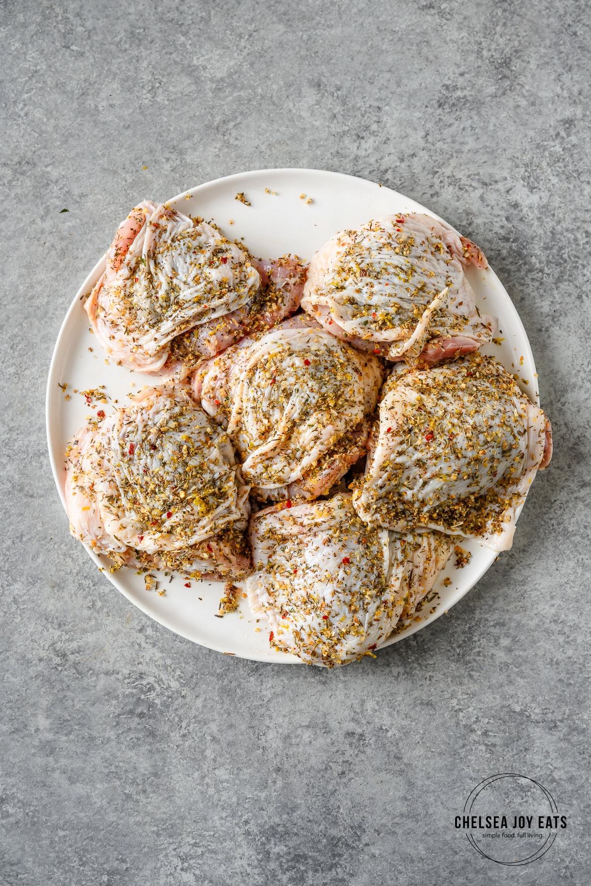 Raw bone-in, skin-on chicken thighs rubbed generously with Greek spices on a white plate.