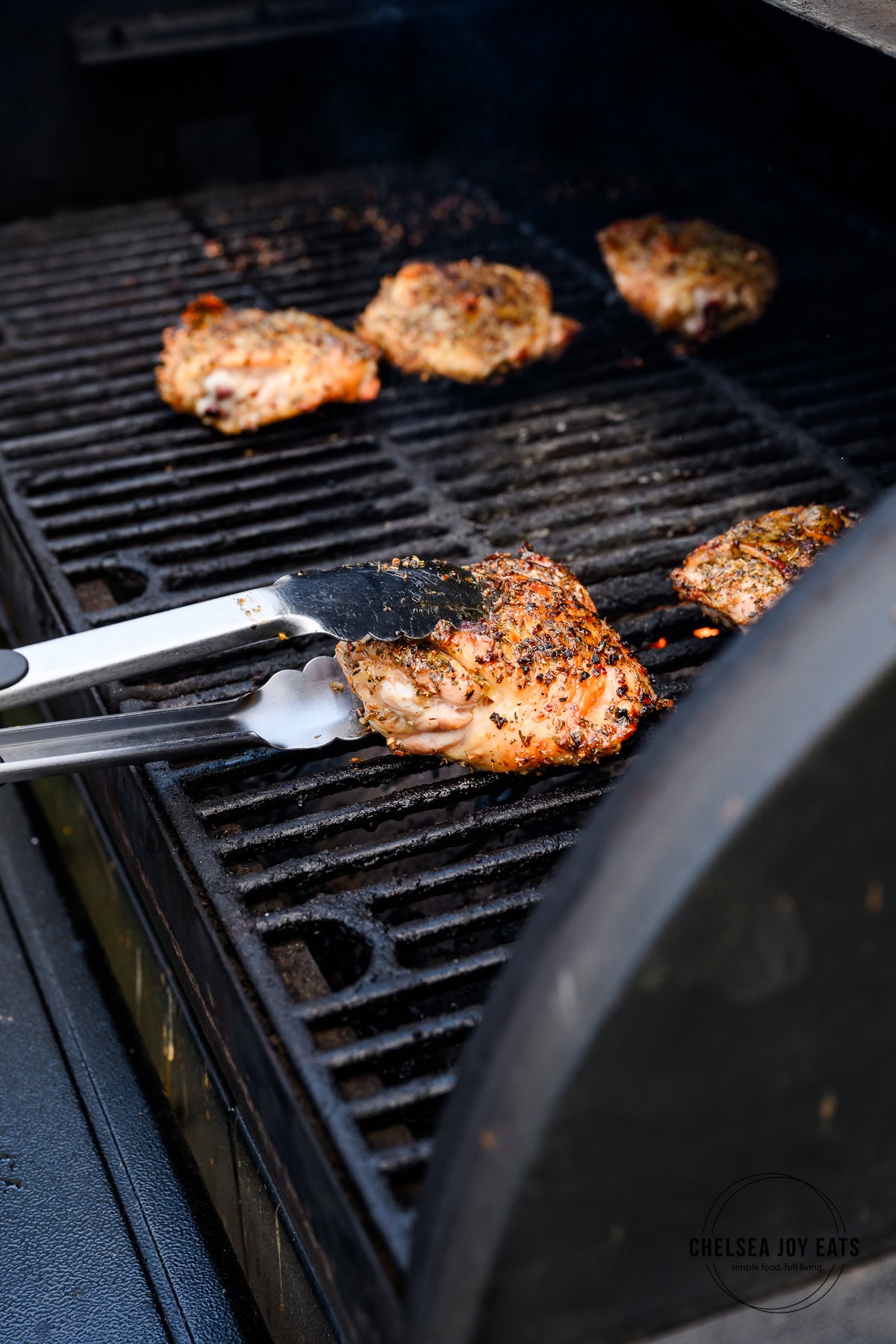 Chicken thighs being turned over by kitchen tongs on a grill to continue cooking.