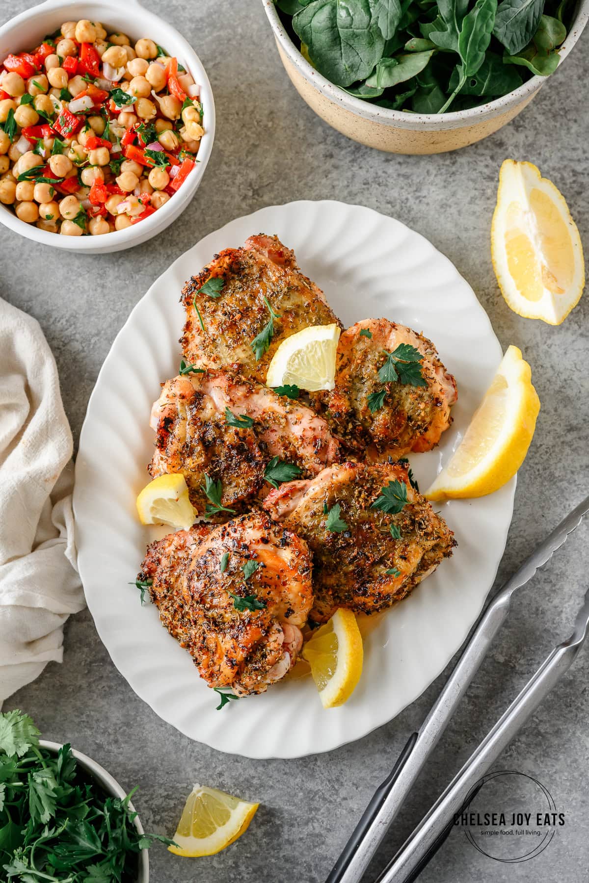 Grilled chicken thighs with Greek seasoning on a serving platter with lemon wedges, with more salad sides nearby.