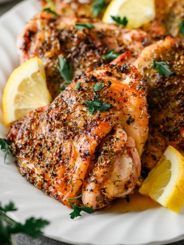 Grilled chicken thigh with Greek seasoning on a serving platter with lemon wedges.