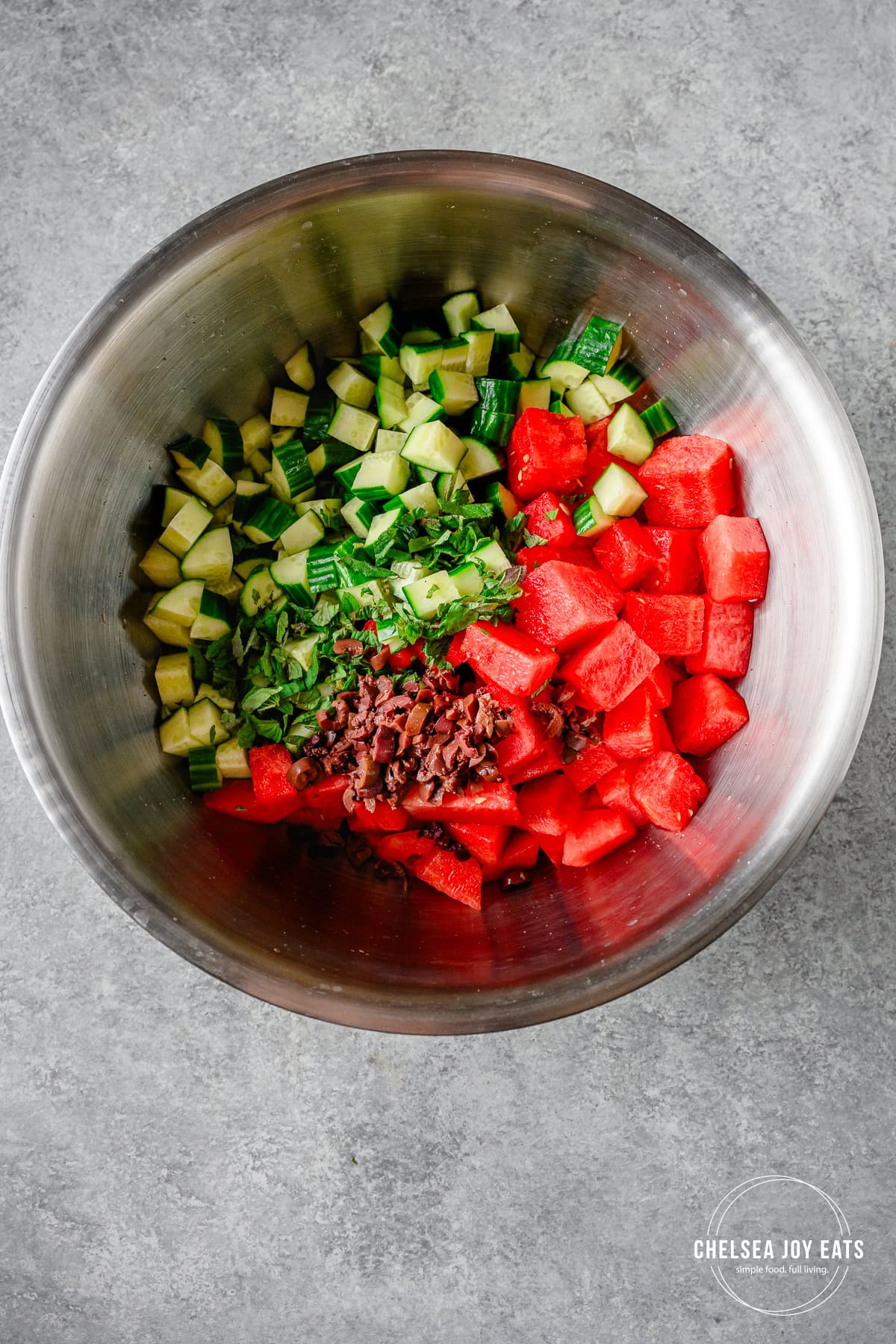 Incredients for a Greek-inspired cucumber and watermelon salad in a large stainless steel bowl.