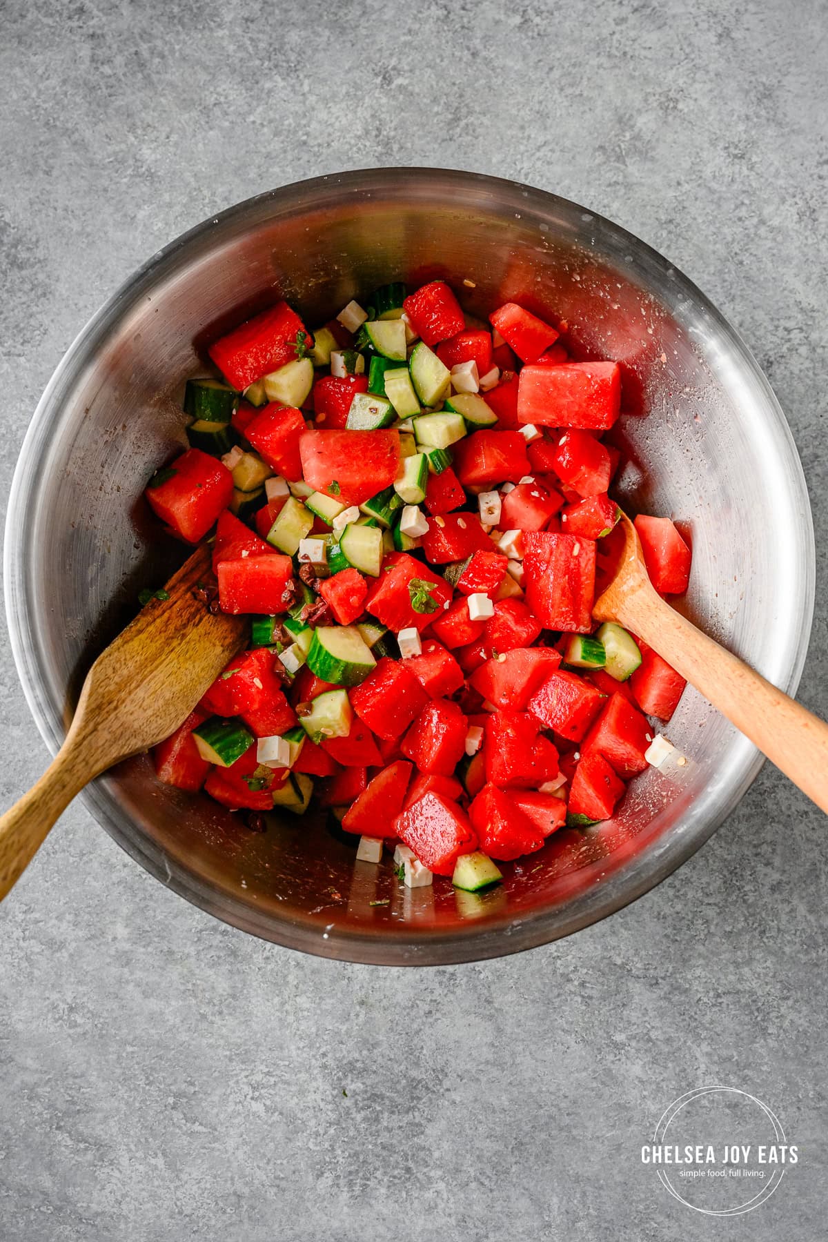 Greek-inspired watermelon and cucumber salad tossed with dressing with wooden spoons in a large stainless steel bowl.