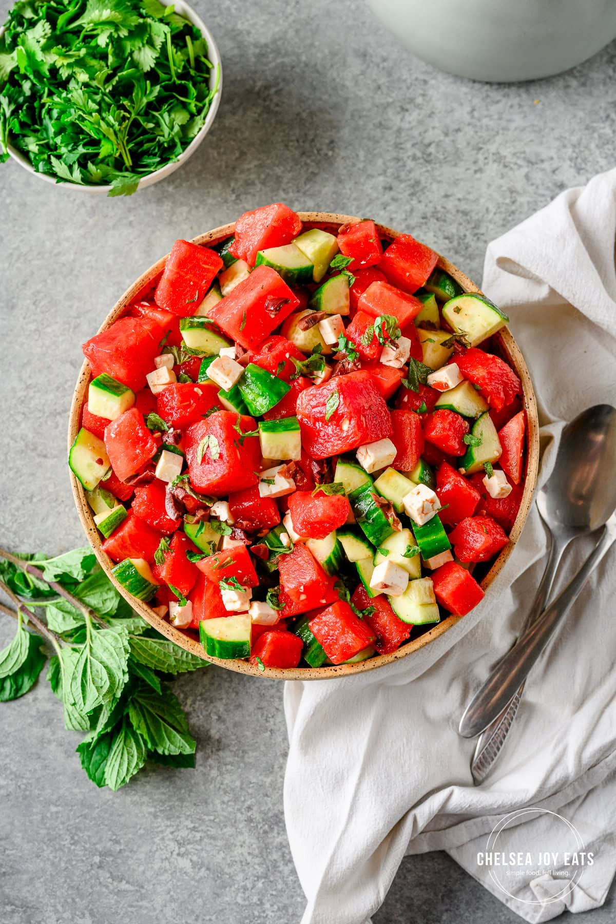 Watermelon and cucumber salad in a large ceramic bowl with a smaller dish of greens, linens, and serving spoons besides.