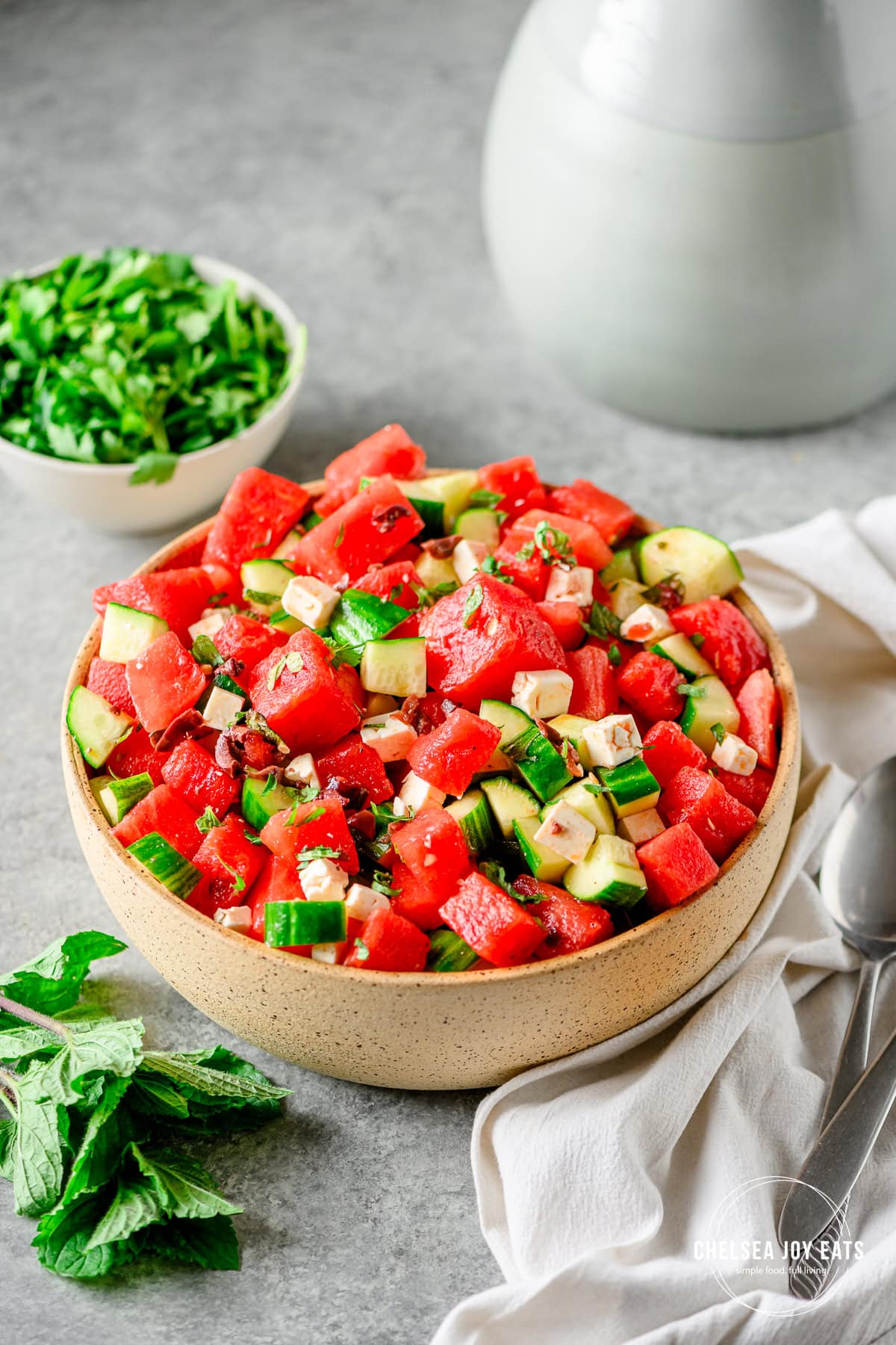 Large ceramic bowl with a Greek-inspired watermelon and cucumber salad with kalamata olives and feta cheese on a light background with linens and serving spoons besides.