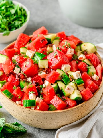 Ceramic bowl with watermelon and cucumber salad along with a small dish of greens in the background.