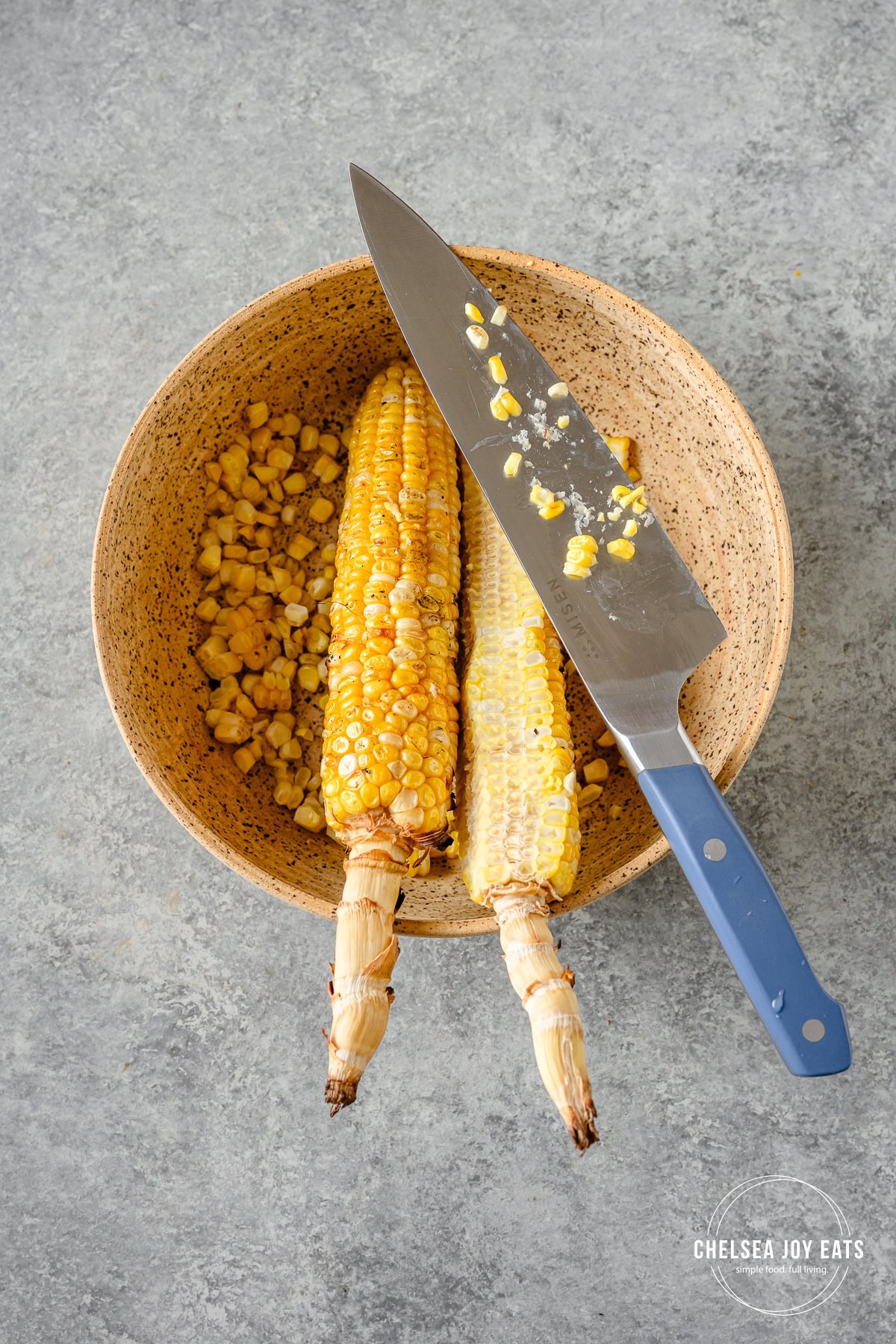 Grilled ears of corn with a knife in a large ceramic bowl, with fresh cut corn kernels in thebowl.