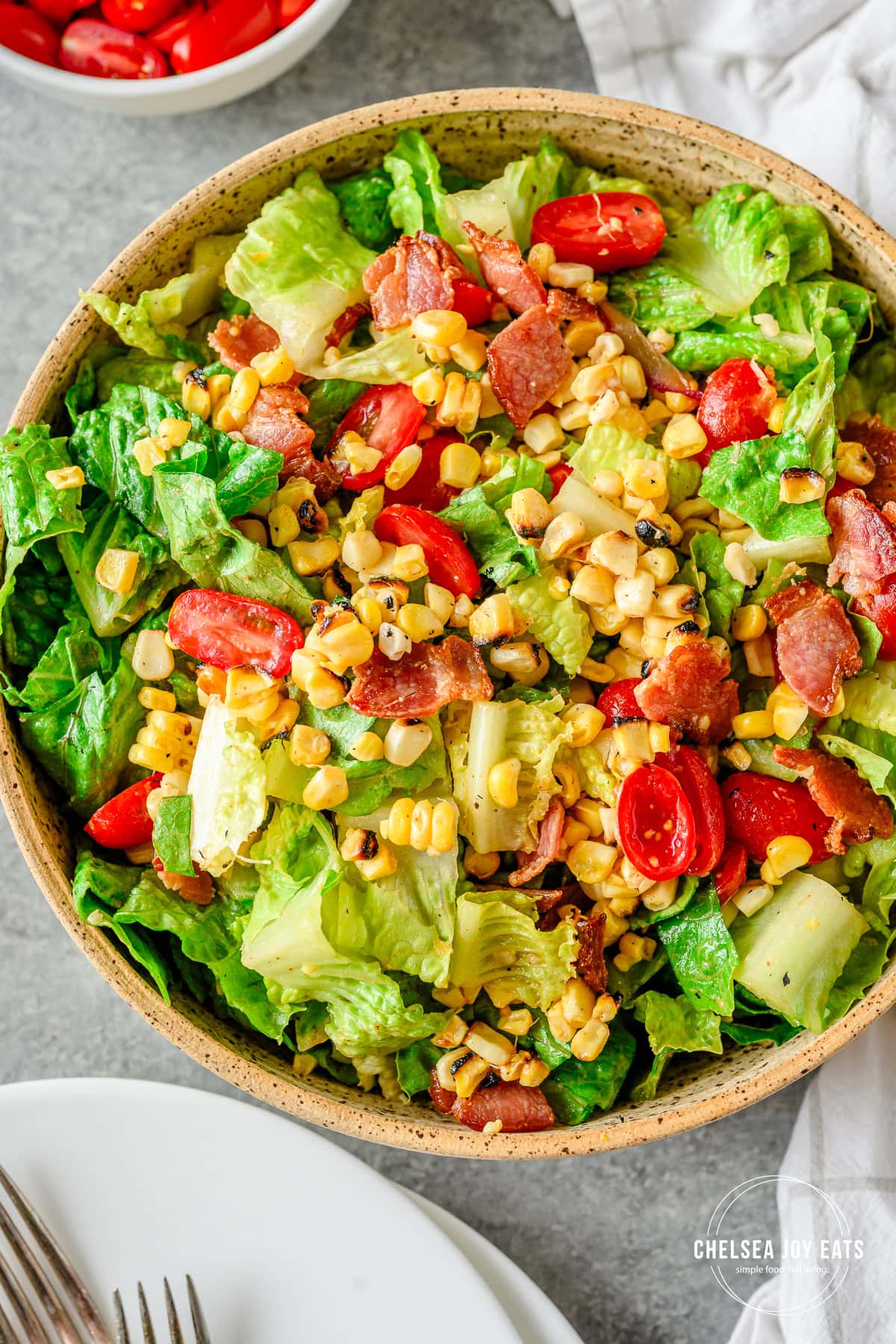 Tossed Corn & Tomato salad with bacon in a large ceramic bowl on a grey countertop with plates and table linens besides.
