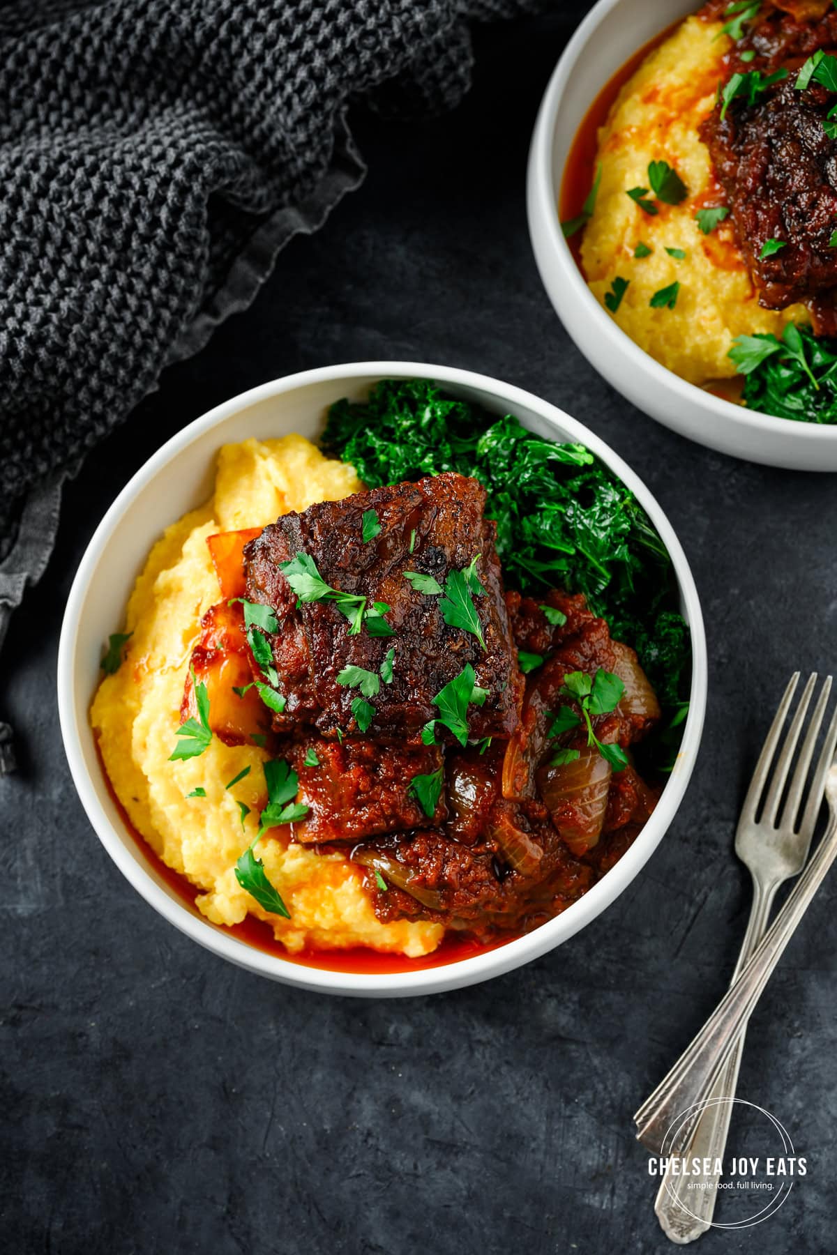 Cooked gluten free balsamic tomato short rib served over polenta with kale in a low bowl with forks alongside.