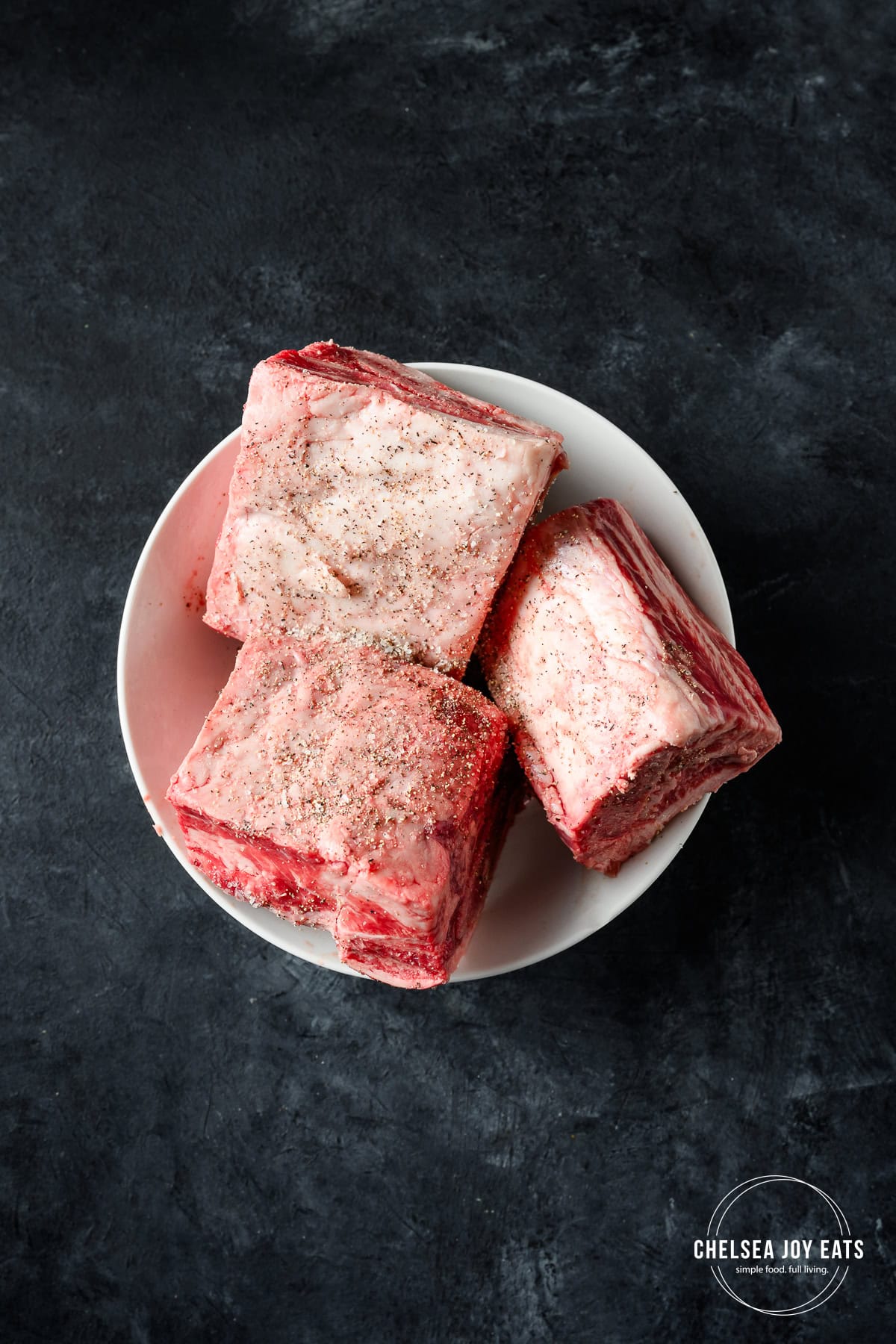 Seasoned uncooked beef short ribs in a bowl against a dark grey countertop.